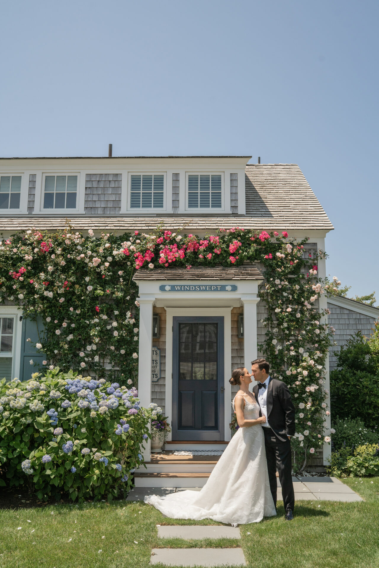 Bride and groom kiss by rose-covered house.