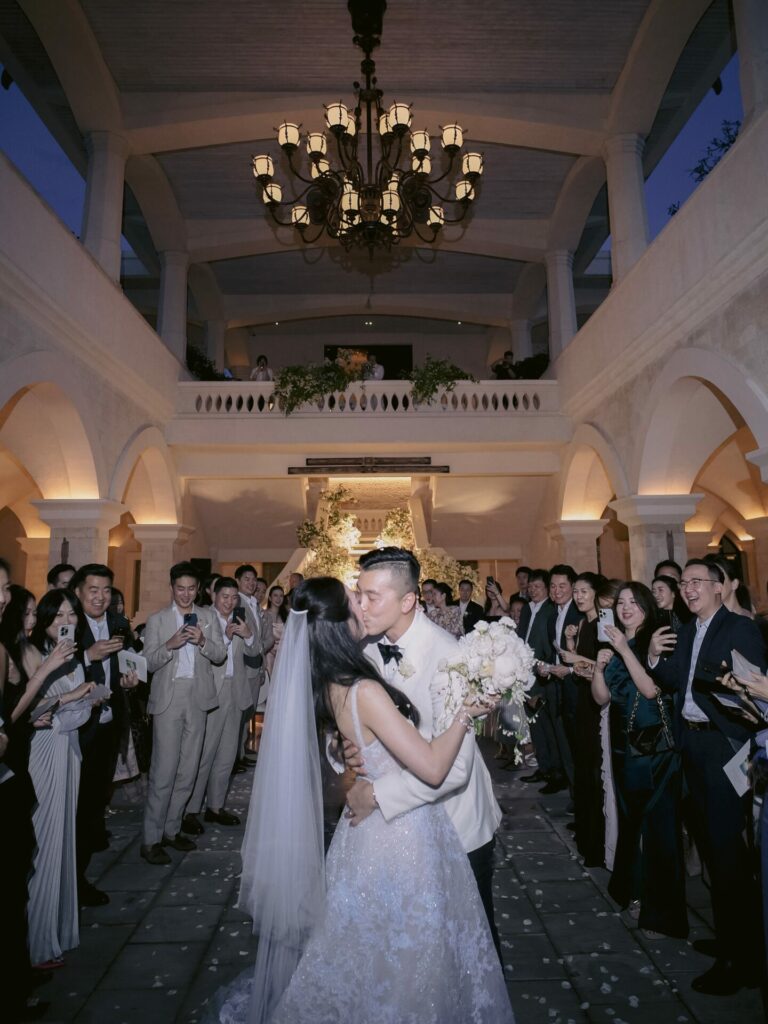 Bride and groom kiss under chandelier, guests cheer.
