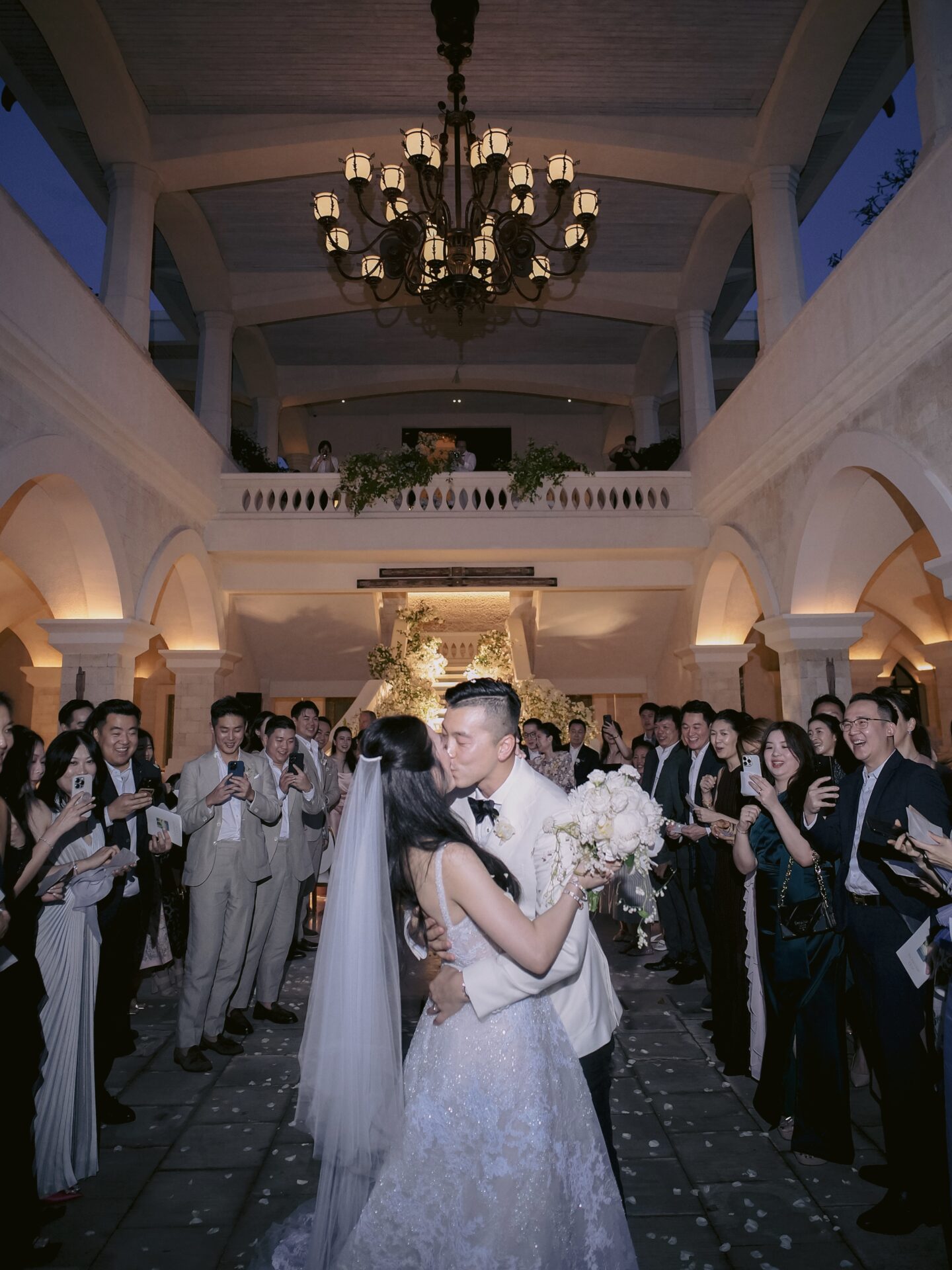 Bride and groom kiss under chandelier, guests cheer.