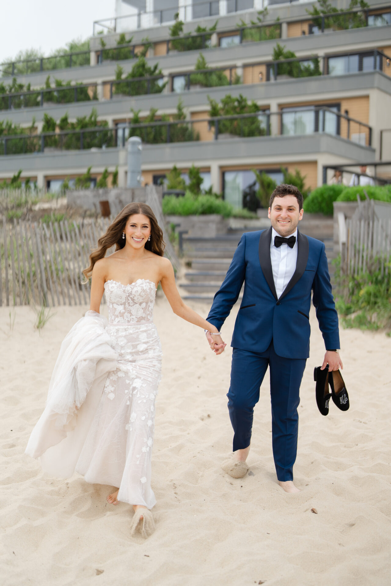 Bride and groom barefoot, smiling on sandy beach.