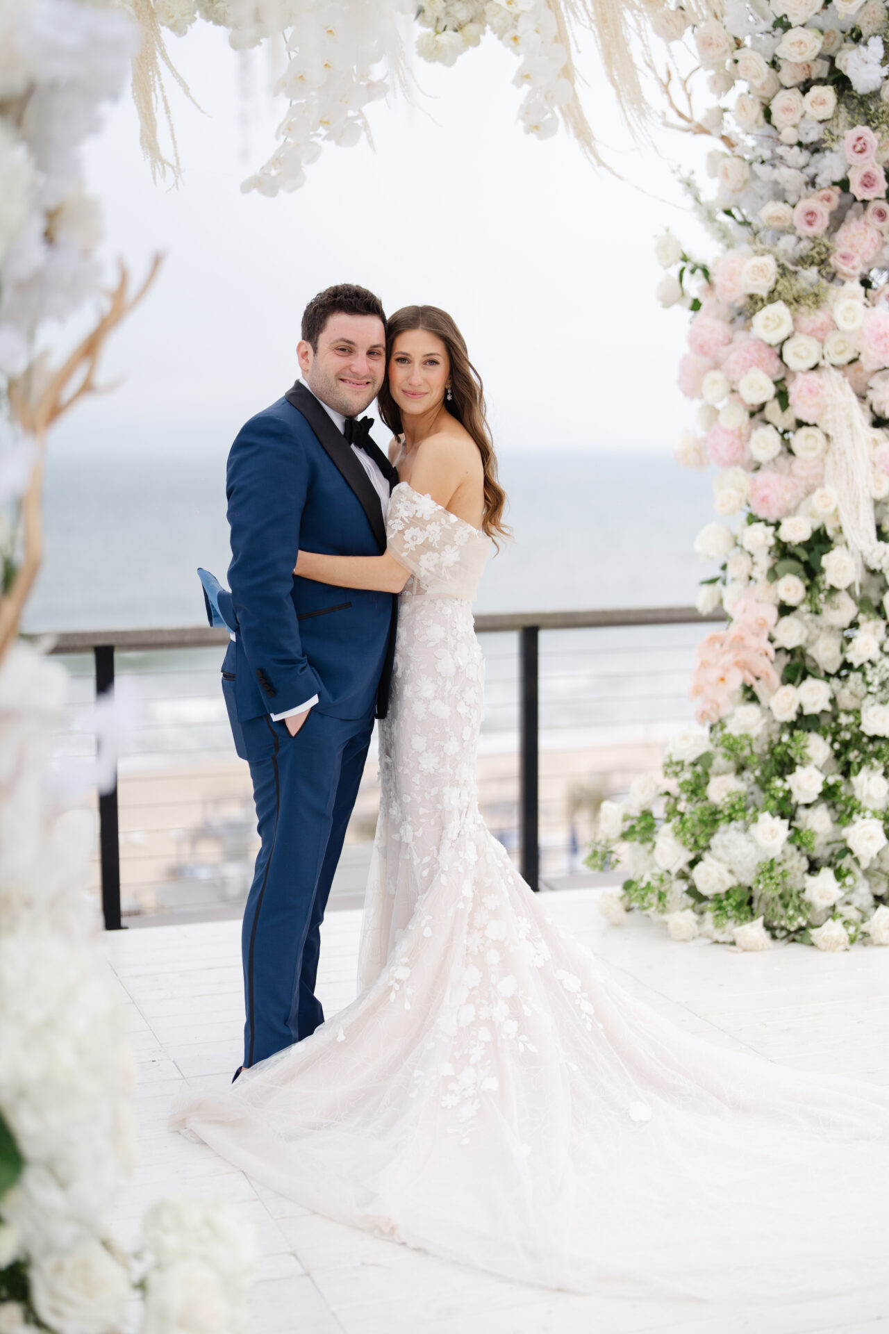 Bride and groom smile under floral arch by ocean.