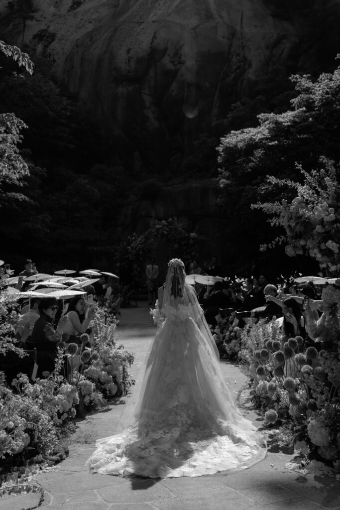 Bride in flowing gown walks outdoor flower aisle.