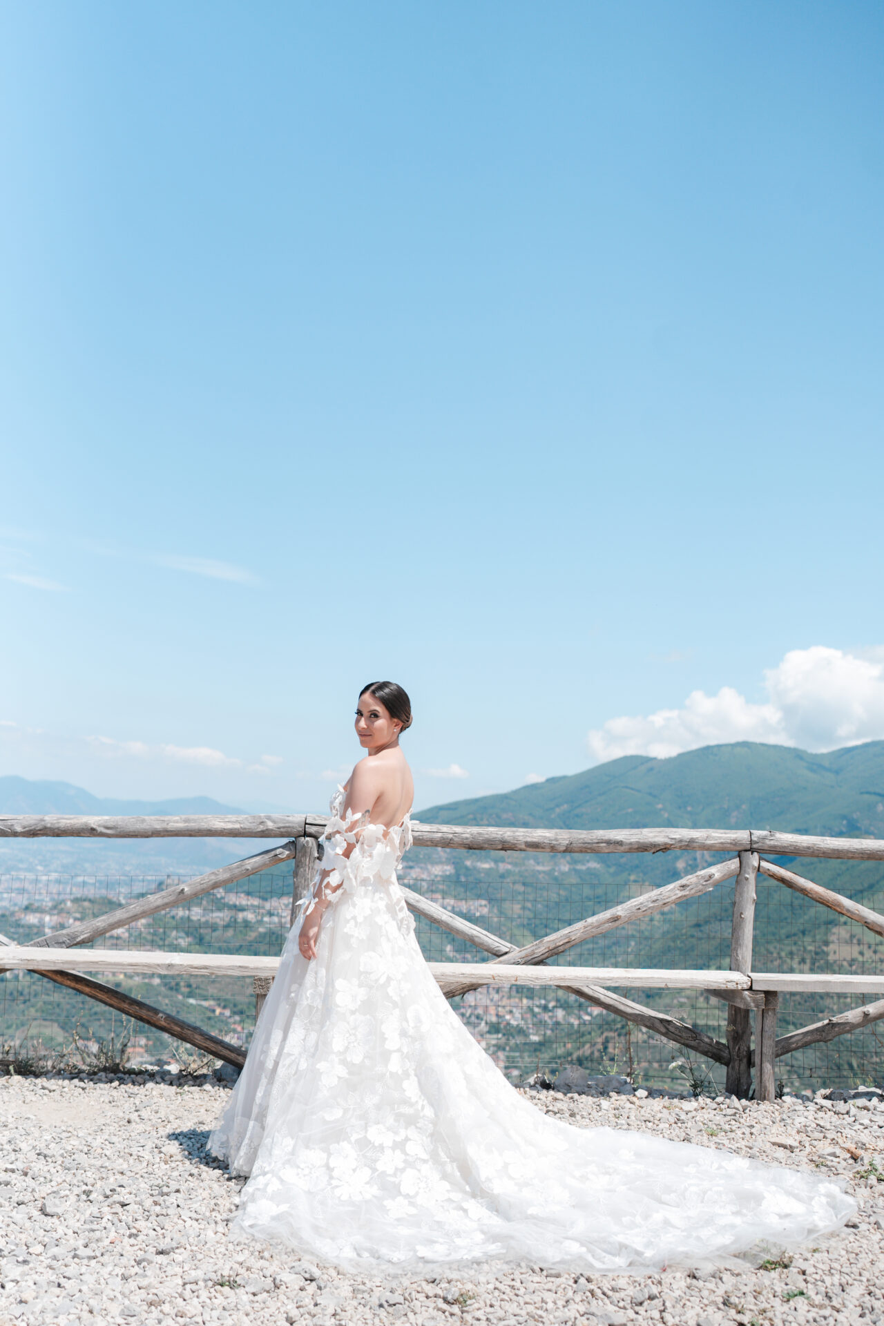 Bride in floral dress by fence, hills behind.