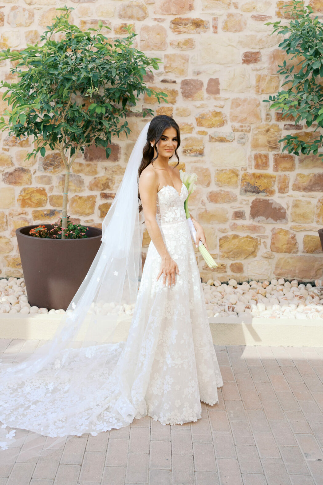 Bride Katya Ruiz in lace gown, bouquet, veiled, framed by potted trees.