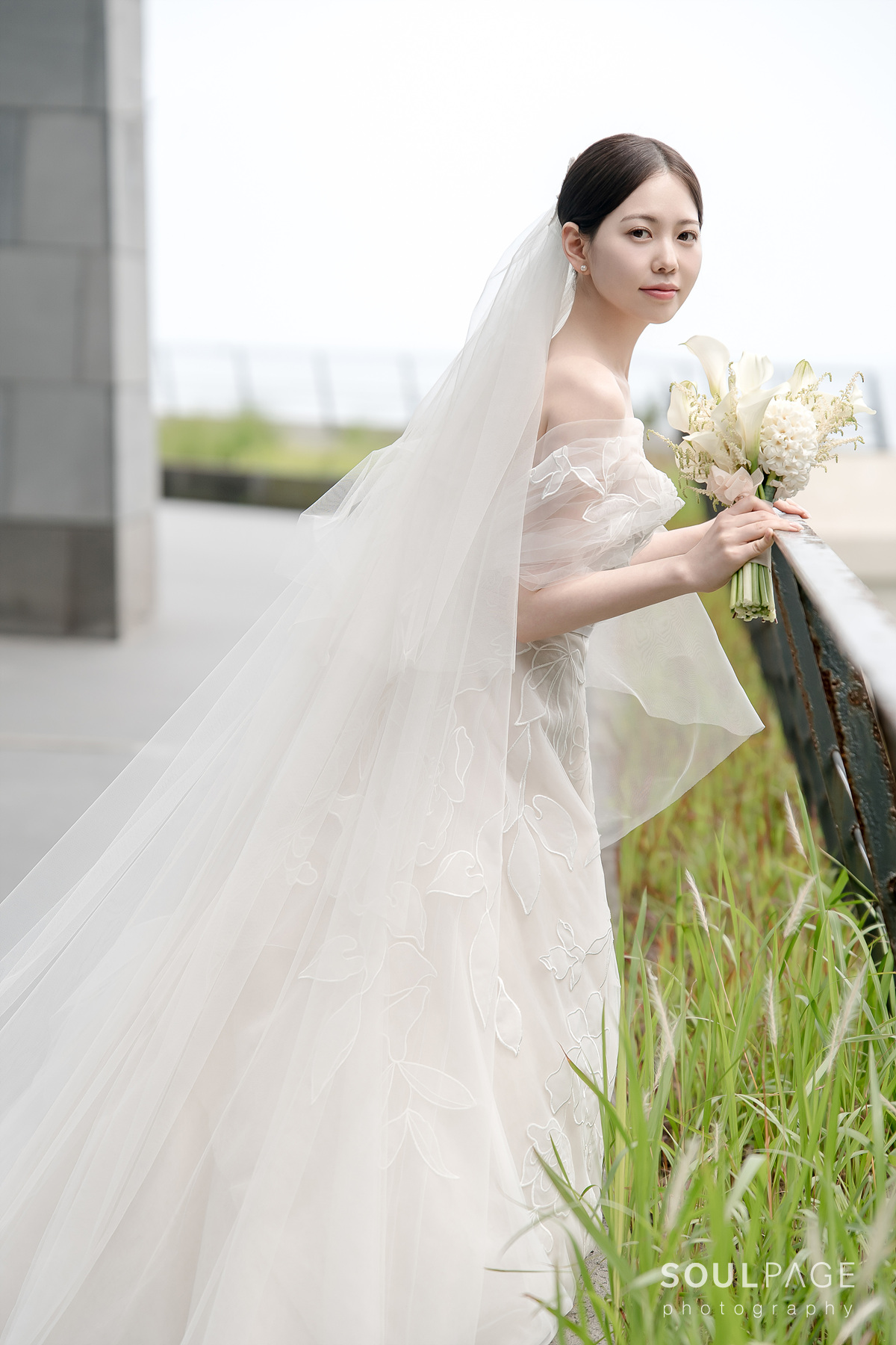 Bride in off-shoulder gown holds white bouquet outdoors.