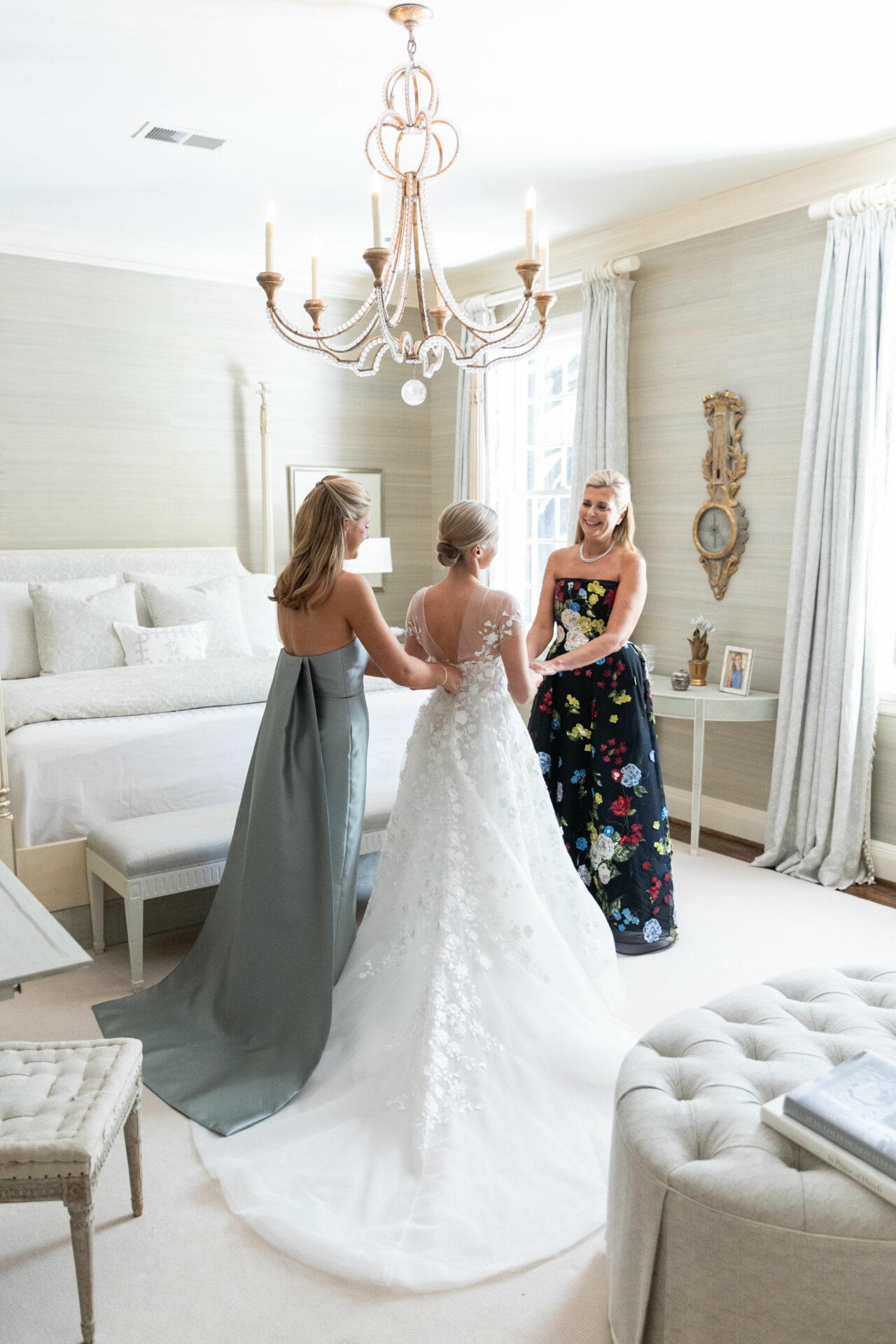 Bride in sunny bedroom, two women adjust dress.