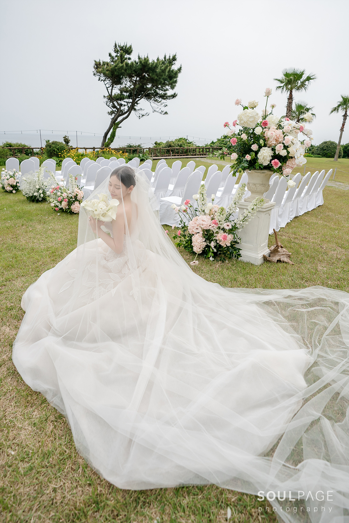 Bride with bouquet sits on grass at outdoor wedding.