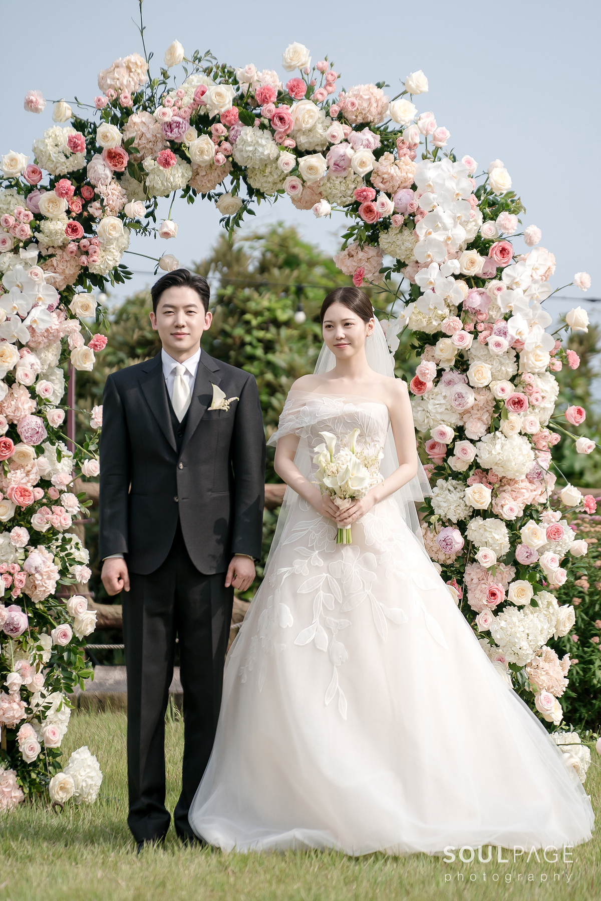 Bride and groom by pink-white floral arch outdoors.