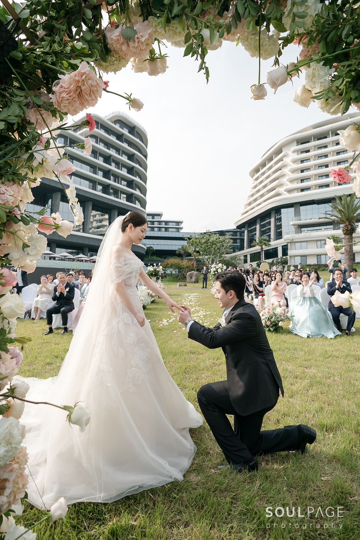 Groom kneels, holding bride’s hand at outdoor wedding.