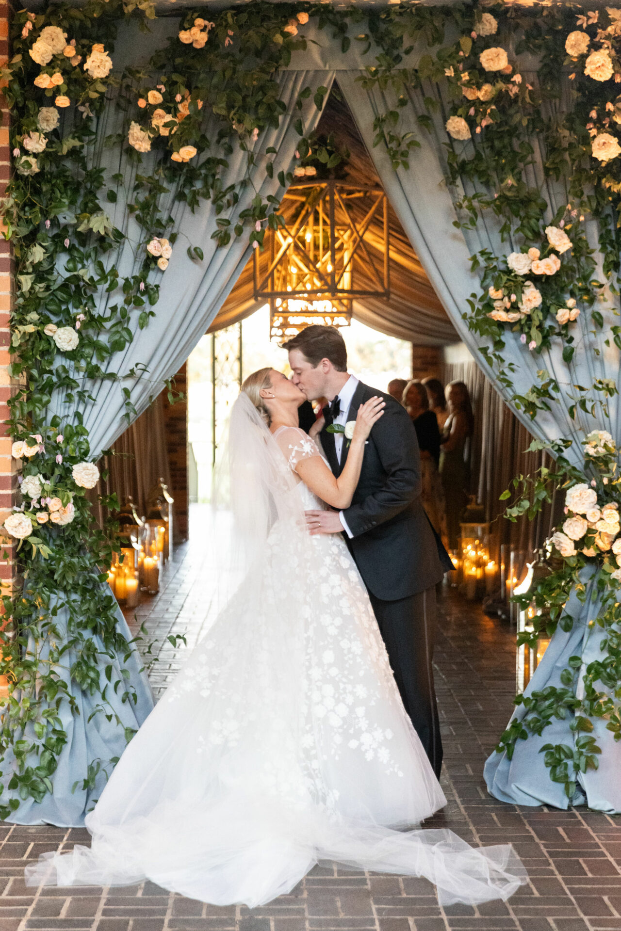Newlyweds kiss under floral arch with blue drapes.