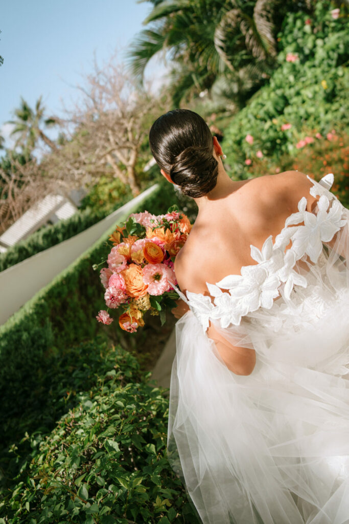 Bride in off-shoulder gown, holding bouquet, walks garden.