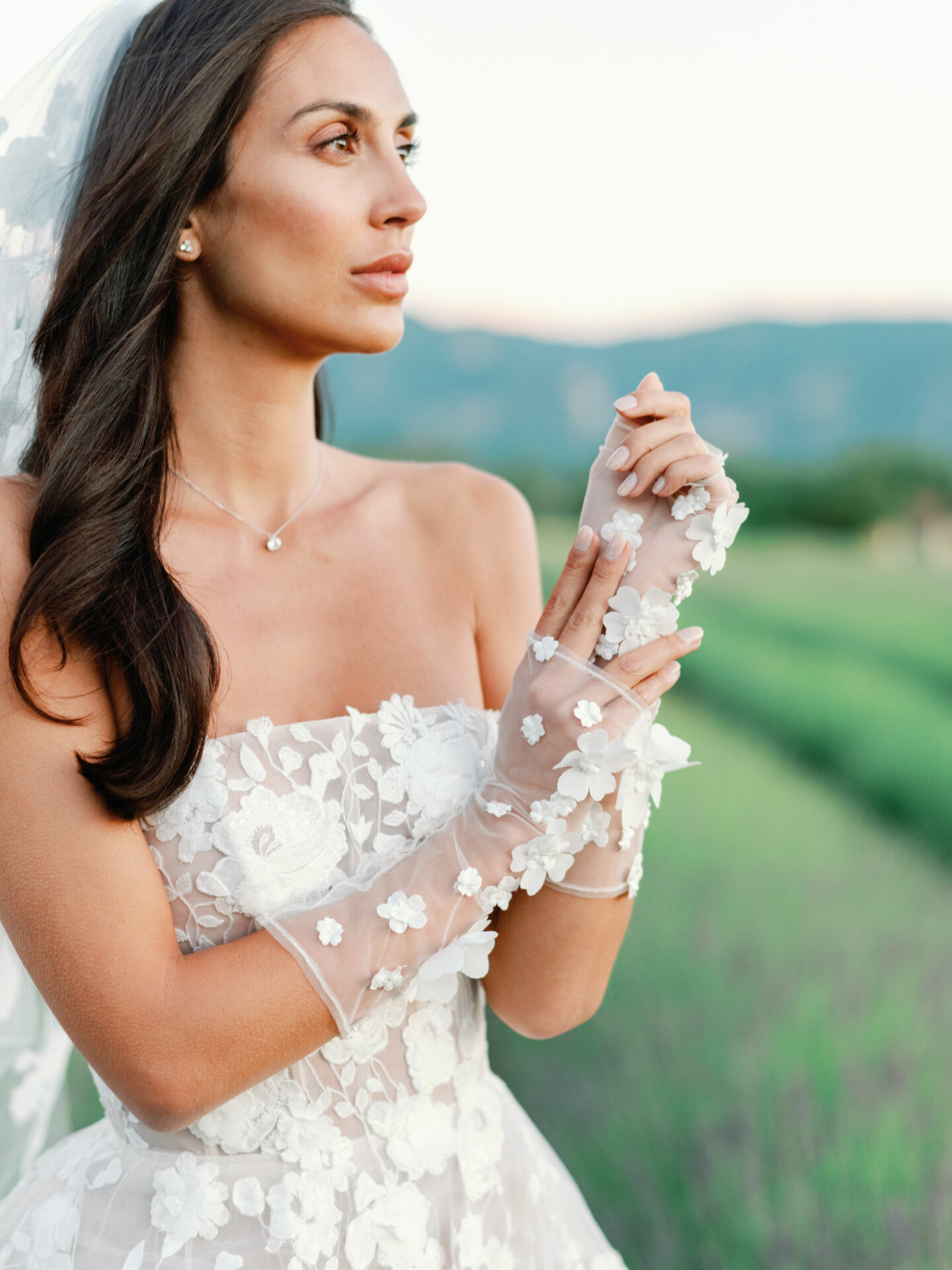 Bride in floral lace dress stands outside, gazing.