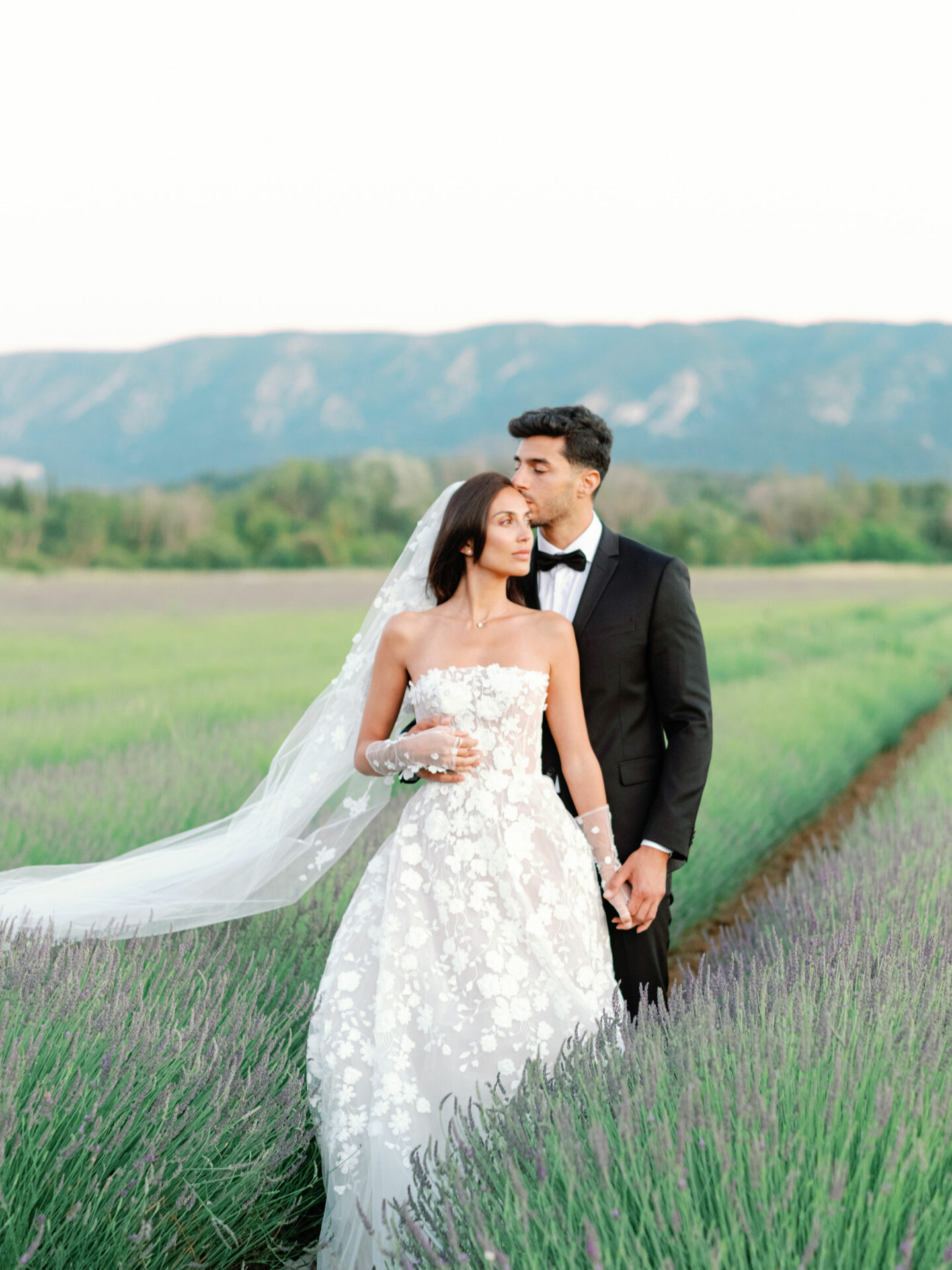 Bride and groom hold hands in lavender field, kissing.