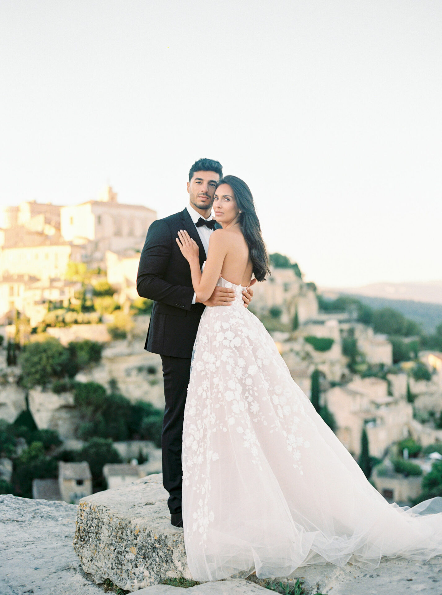 Bride and groom embrace on rocky ledge, village behind.
