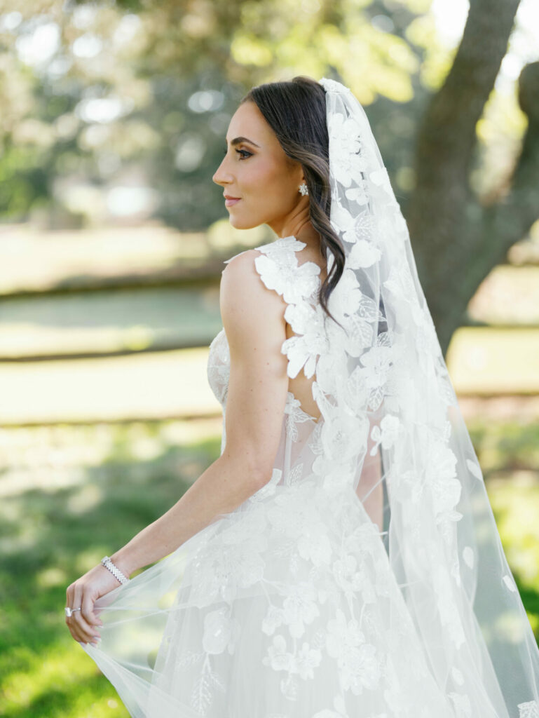 Bride in lace dress and veil stands outdoors.