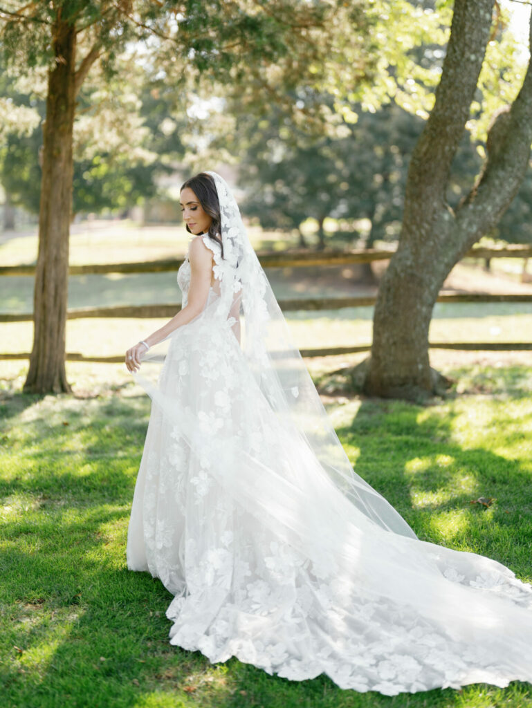 Bride in lace dress with veil stands outdoors.