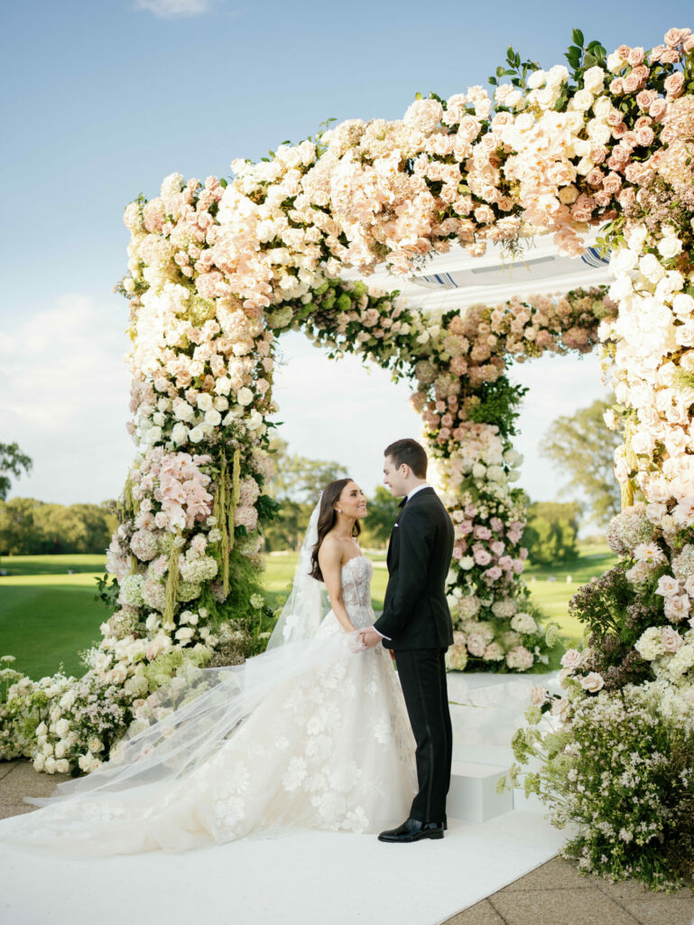 Bride and groom hold hands beneath outdoor flower arch.