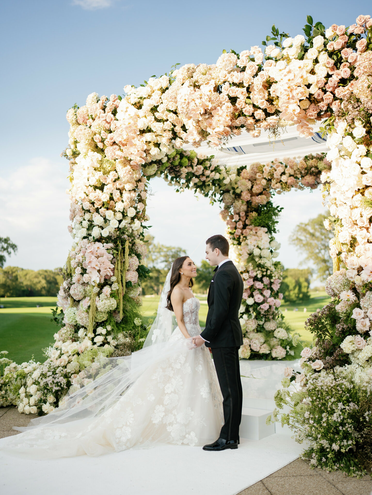 Bride and groom hold hands beneath outdoor flower arch.
