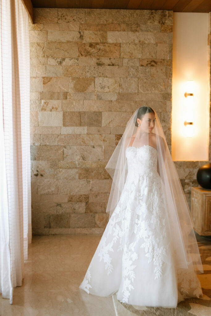 Bride in lace gown stands by stone wall indoors.