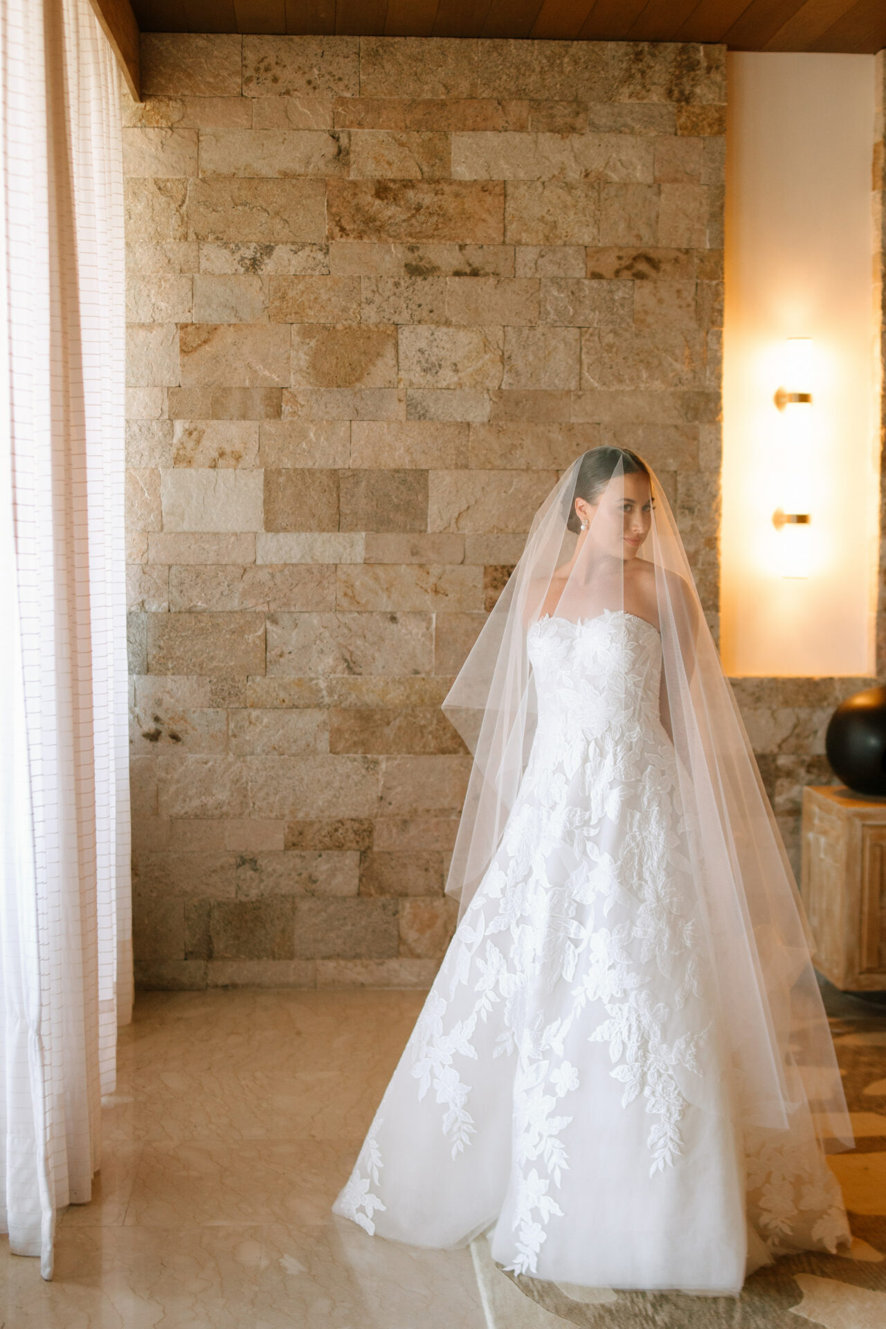 Bride in lace gown stands by stone wall indoors.