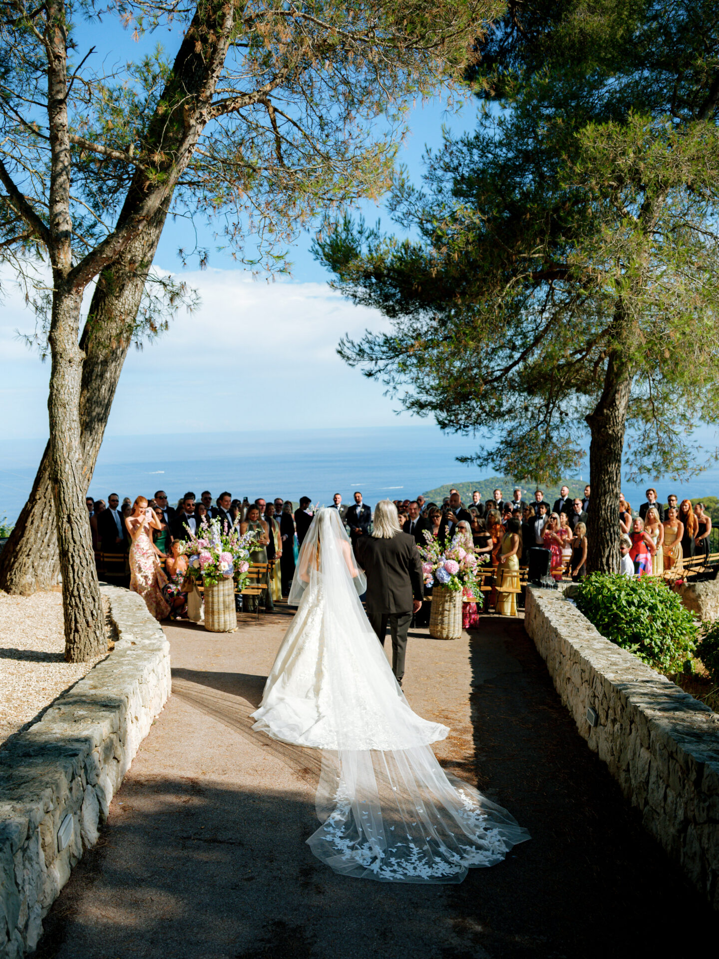 Bride and older man walking outdoor aisle, ocean behind.