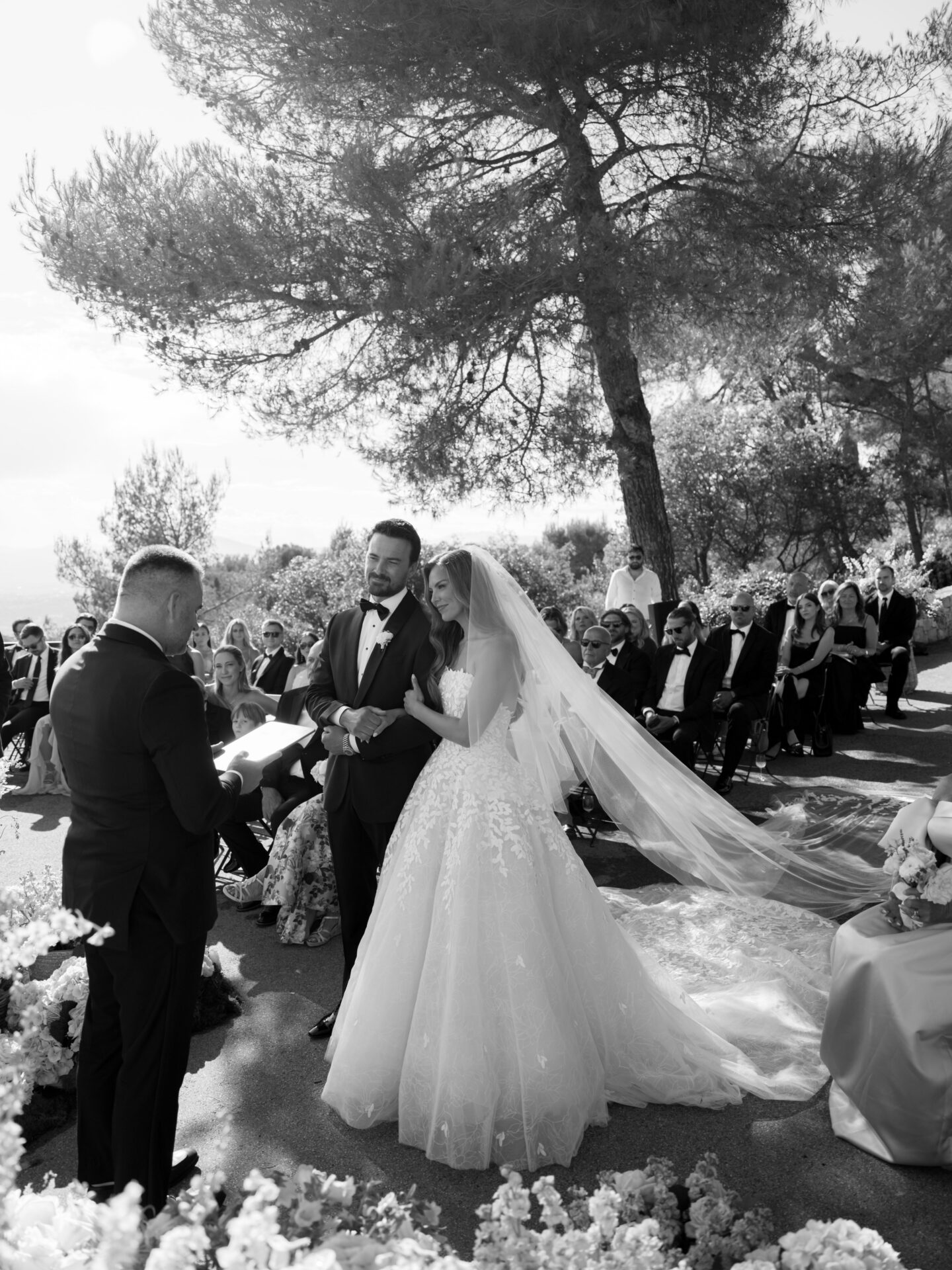 Bride and groom hold hands outdoors, guests seated, sunlight.