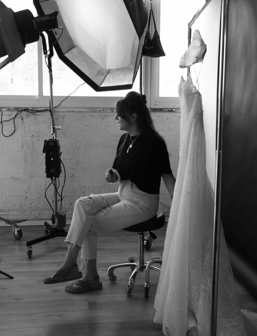 Woman seated in studio beside wedding dress, monochrome.