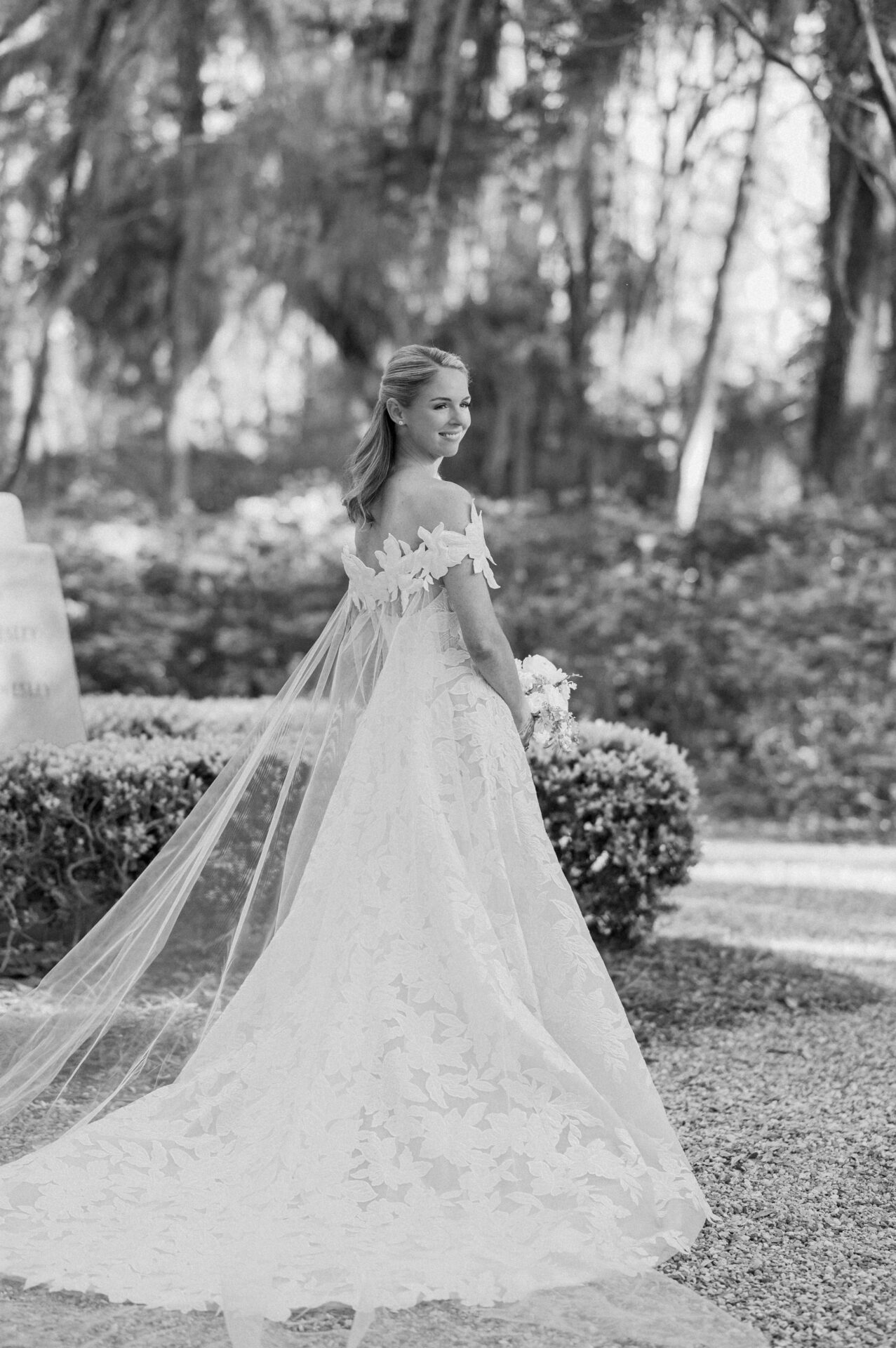 Bride smiles outdoors in lace gown, holding bouquet.