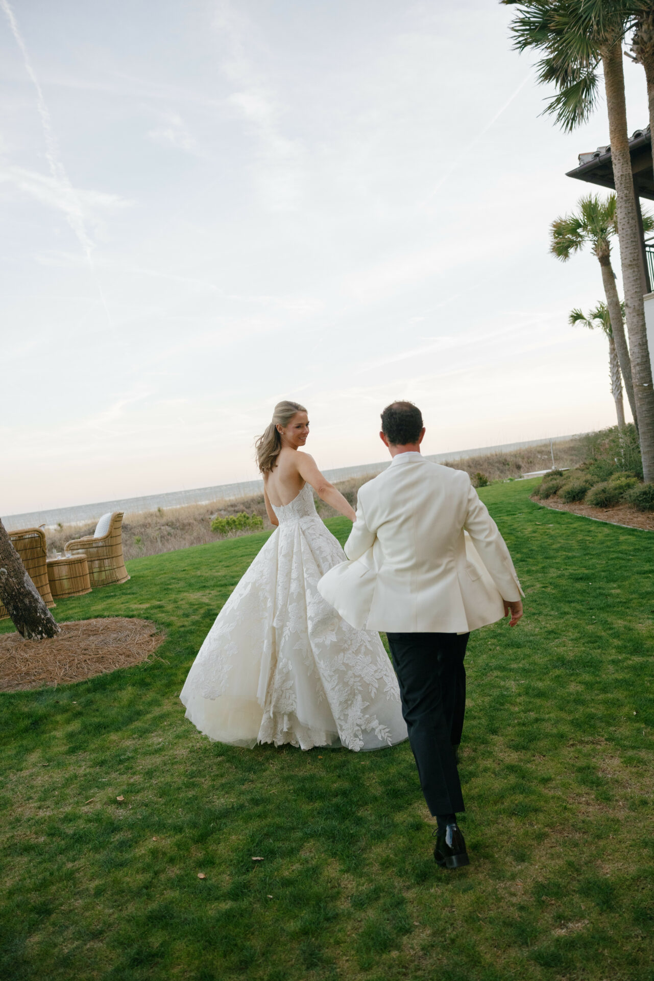 Bride and groom walk hand-in-hand by palms, ocean.