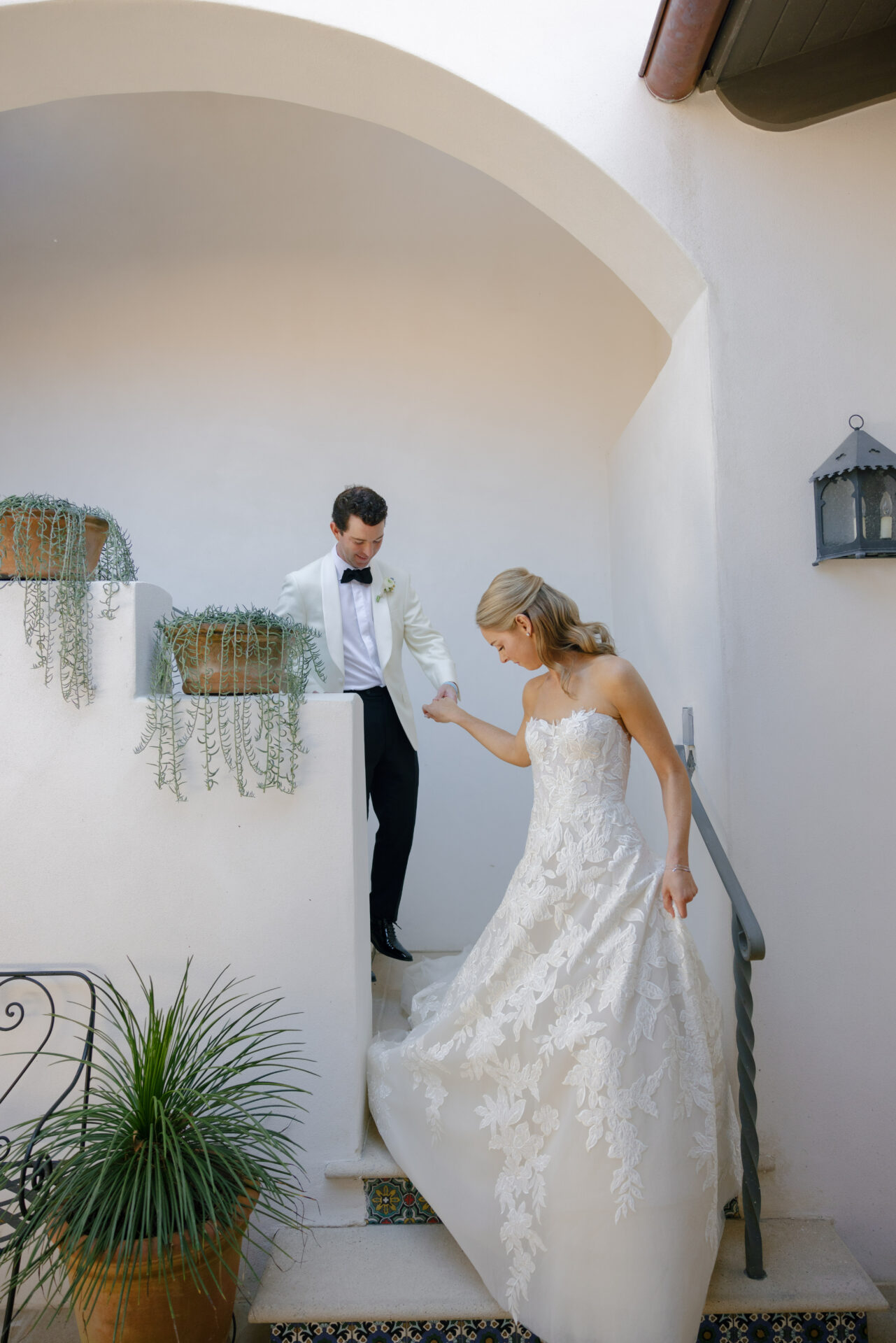 Bride and groom descend outdoor stairs, plants around.