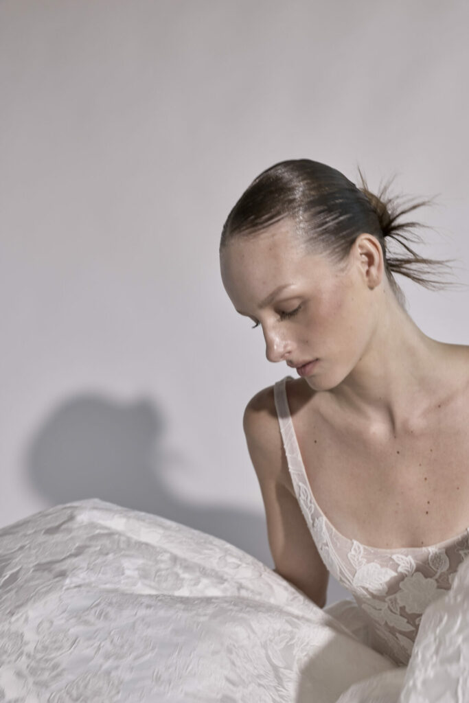 Woman in white lace gown looks down, softly lit.