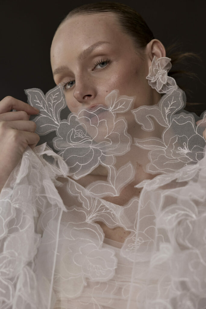 Woman in sheer white lace gown, hand near face.