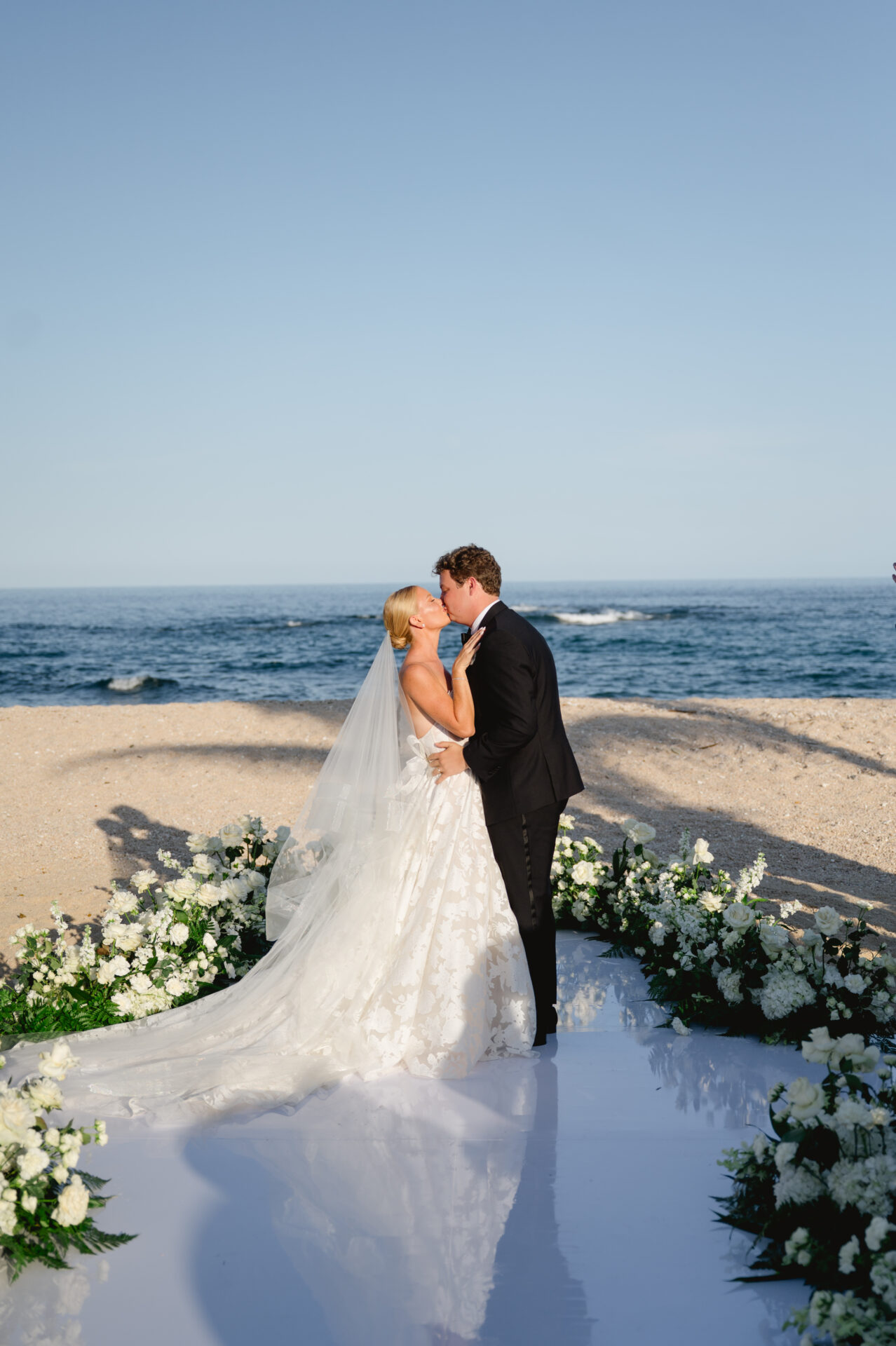 Bride and groom kiss on beach with flowers.