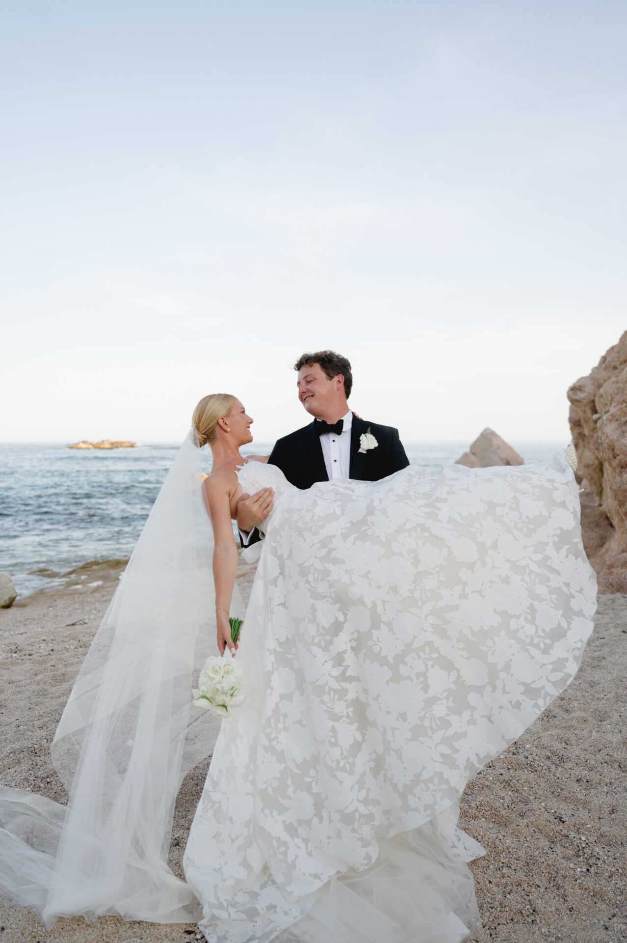 Smiling bride and groom embrace on rocky beach.