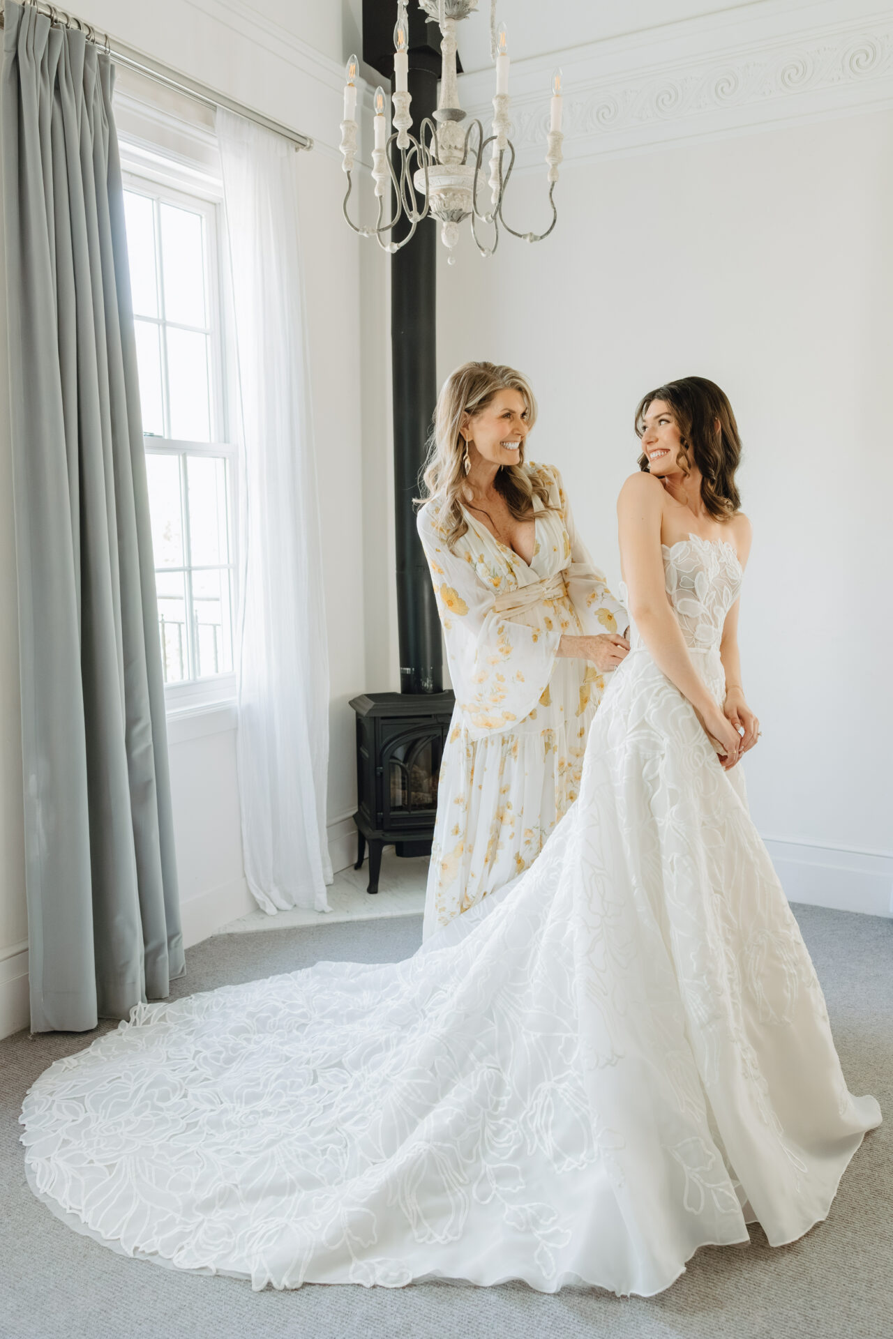 Bride in white gown smiles as woman assists.