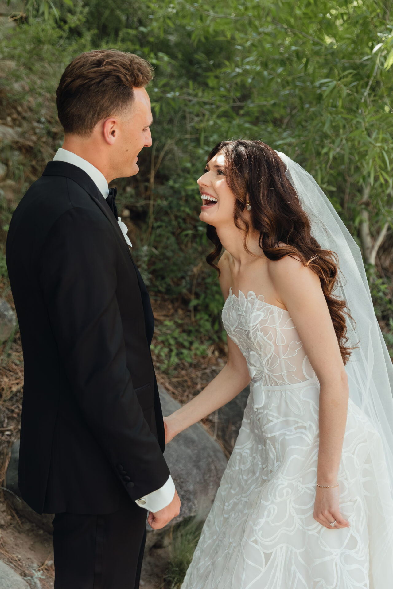Bride and groom laughing outdoors among trees and rocks.
