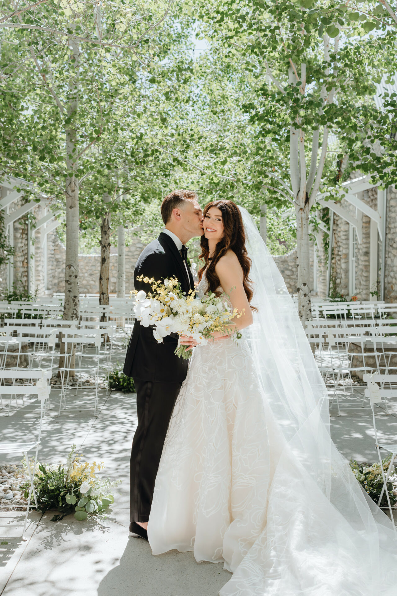 Groom kisses smiling bride with bouquet under leafy trees.