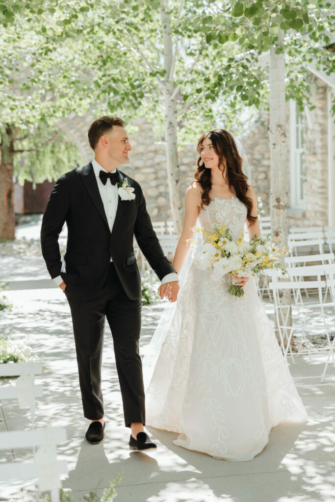 Bride and groom walk outdoors, smiling, holding hands.