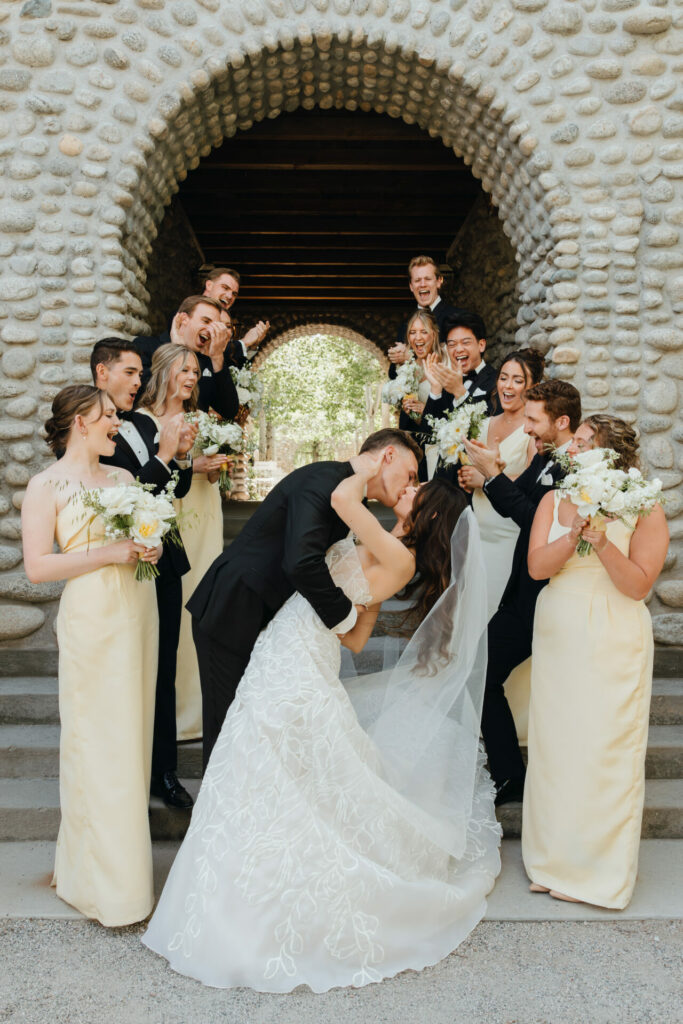 Bride and groom kiss, dipped amid smiling wedding party.