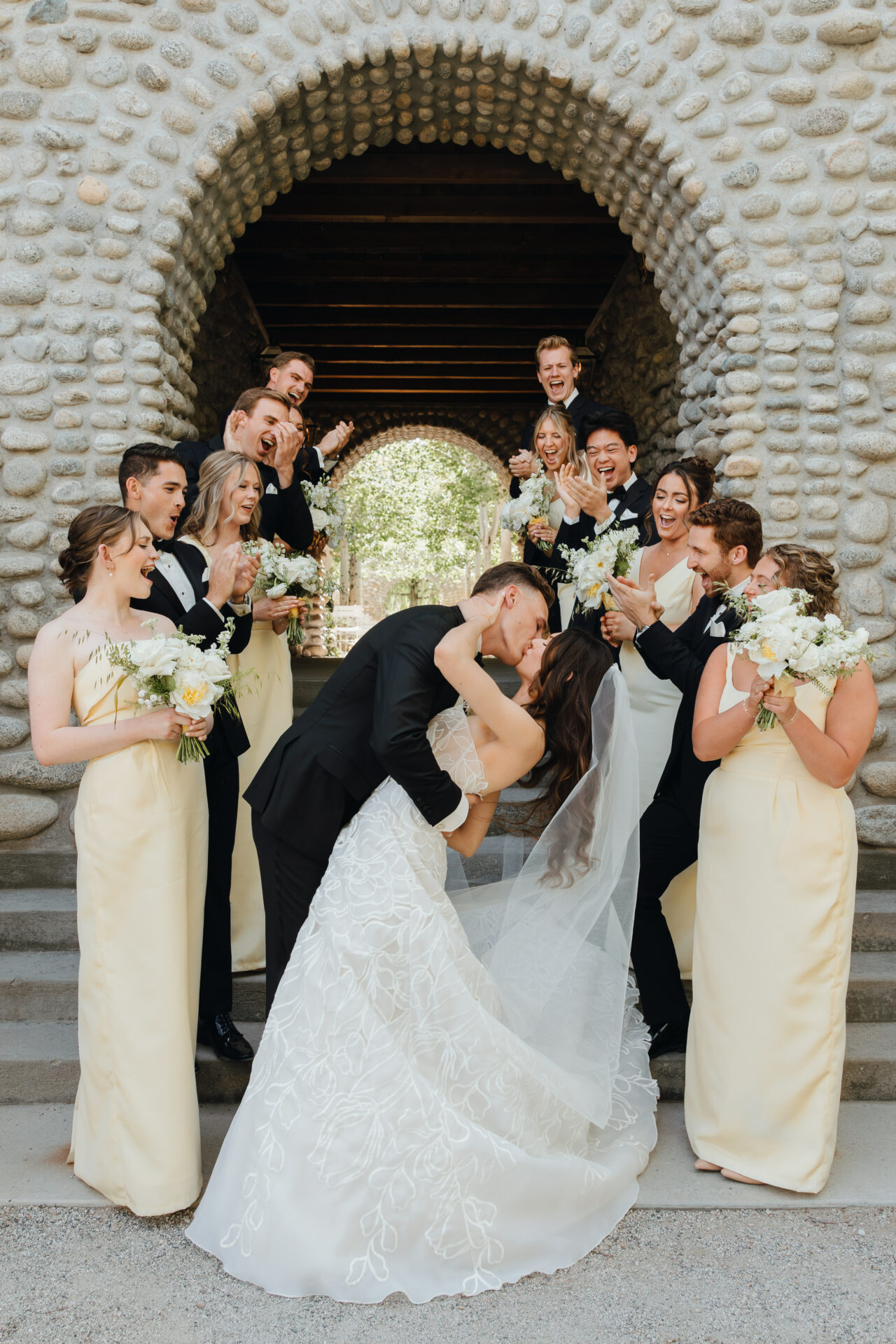 Bride and groom kiss, dipped amid smiling wedding party.