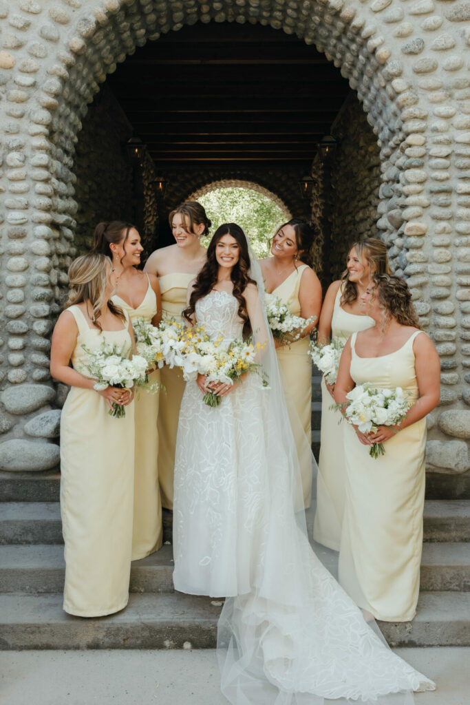 Bride and five bridesmaids in yellow, under stone arch.