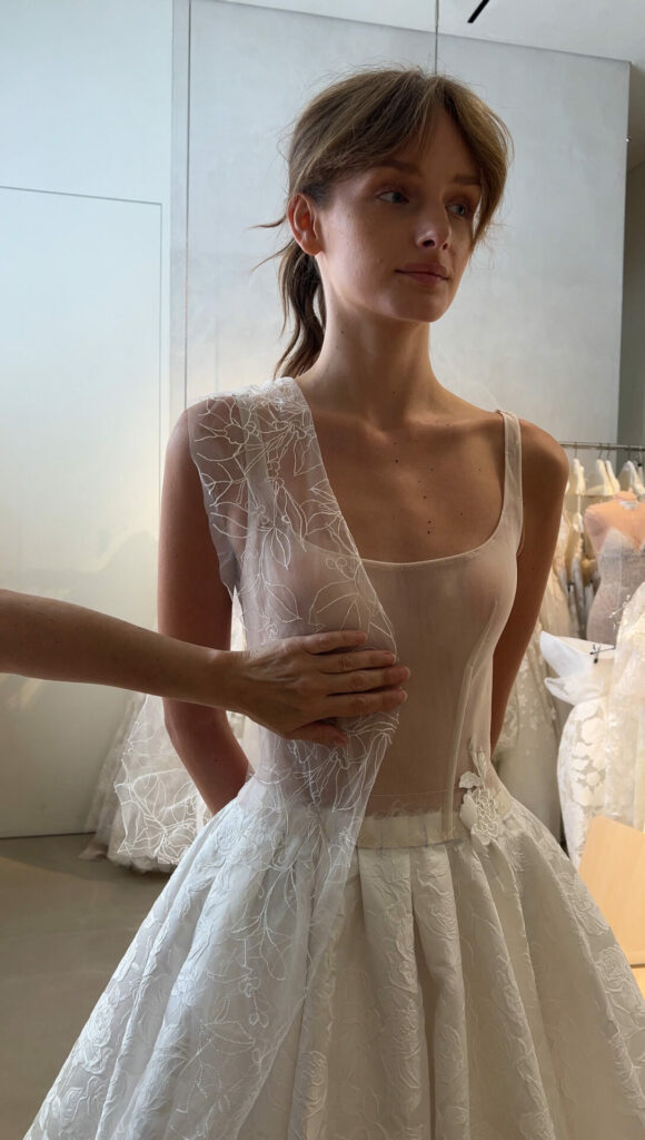 Woman fitted in sheer white dress, ball gowns behind.