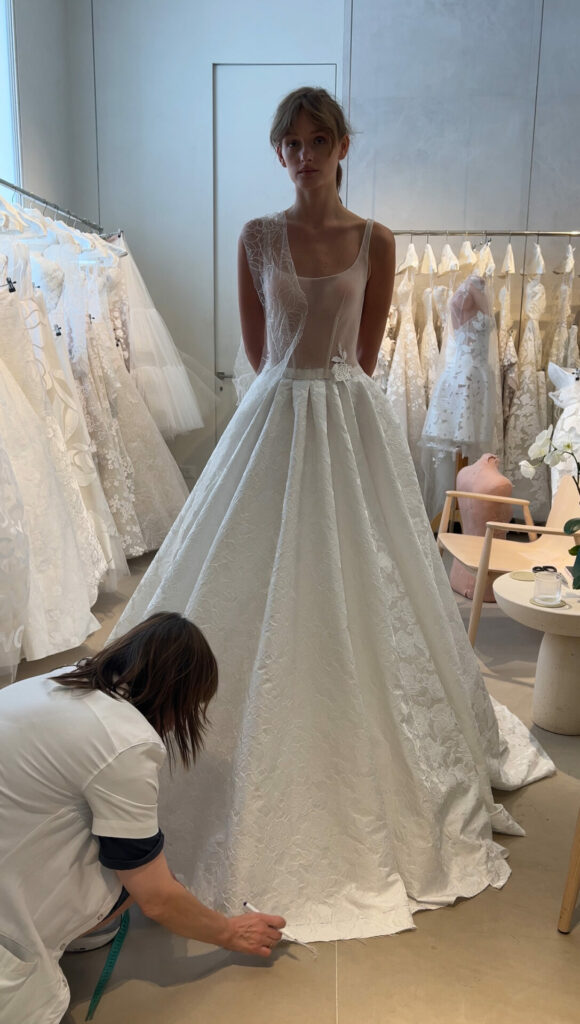 Bride in sheer gown fitted by tailor in boutique.
