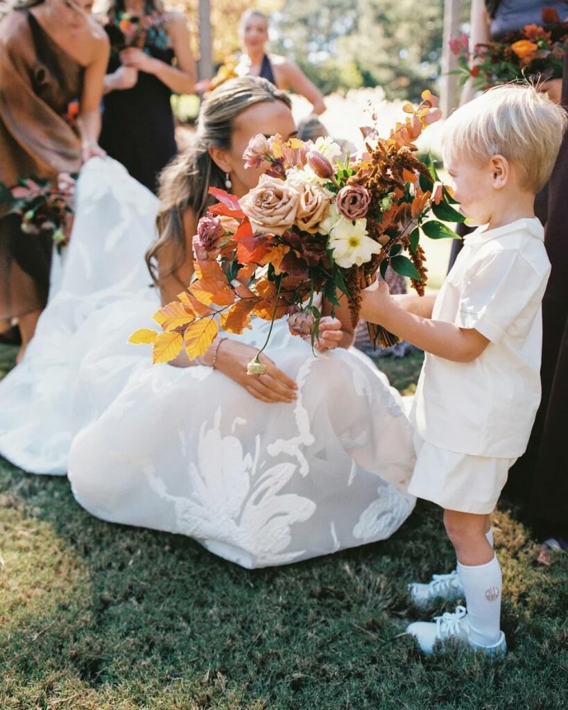 Bride kneels on grass with bouquet, boy beside; bridesmaids behind.