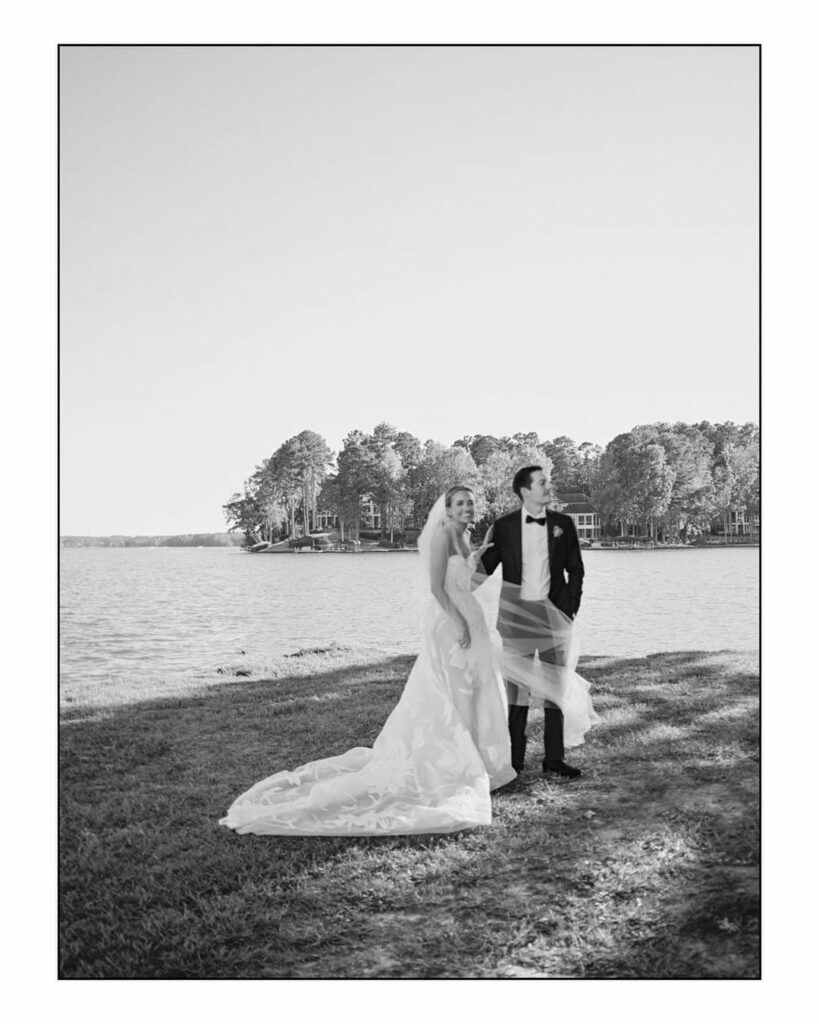 Bride and groom by lake, gown flowing, black and white.