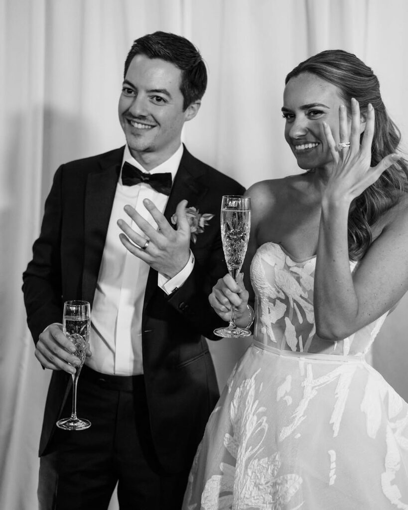 Smiling newlyweds in luxe attire show rings and toast.