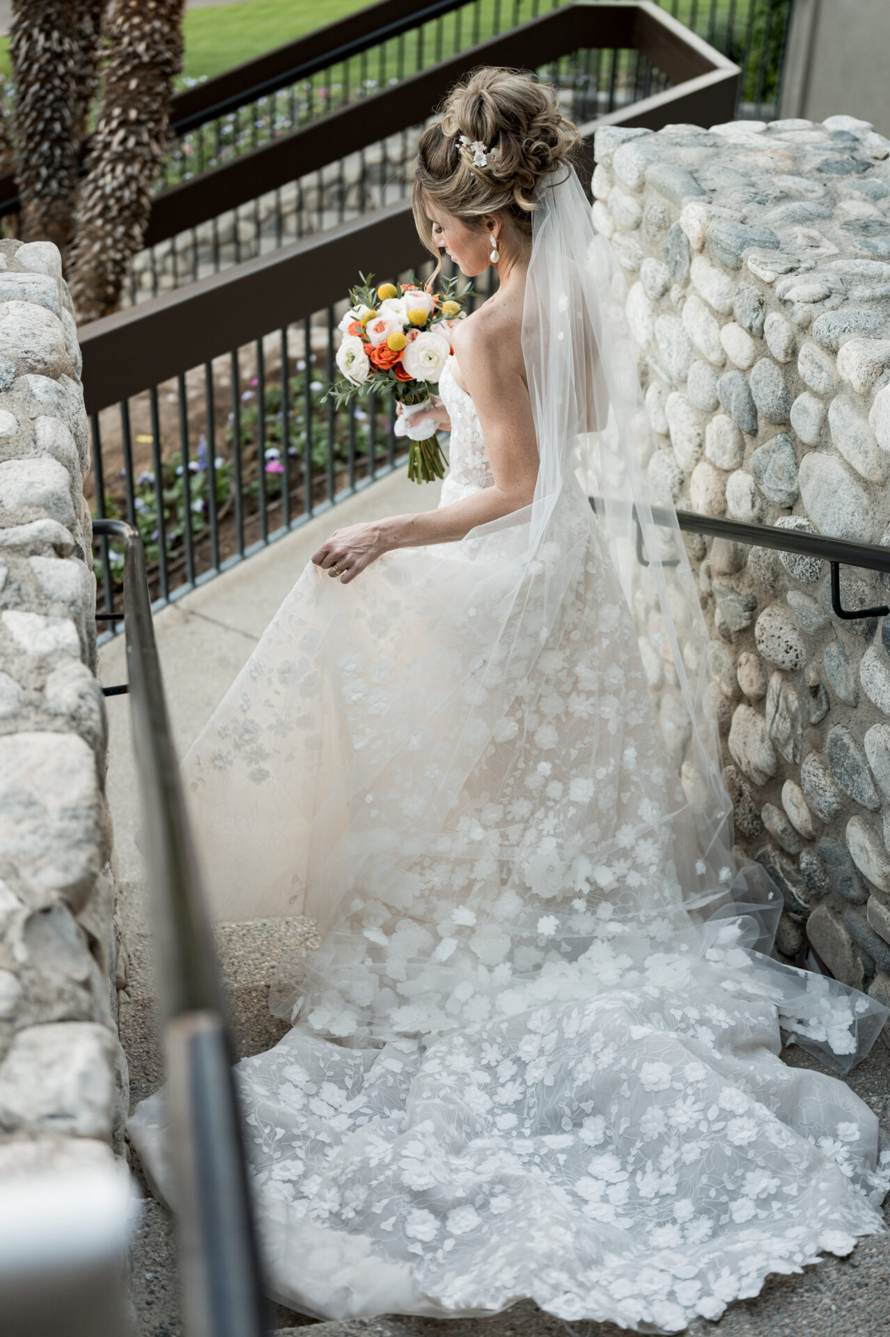 Bride in lace gown climbs outdoor stone steps with bouquet.