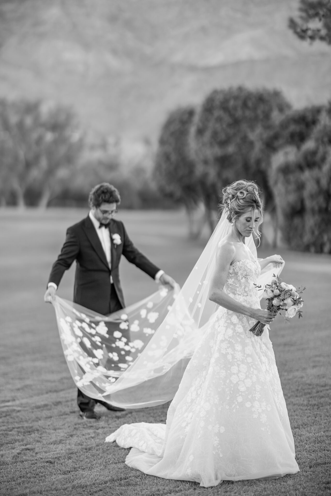 Bride with bouquet, groom arranges veil outdoors.