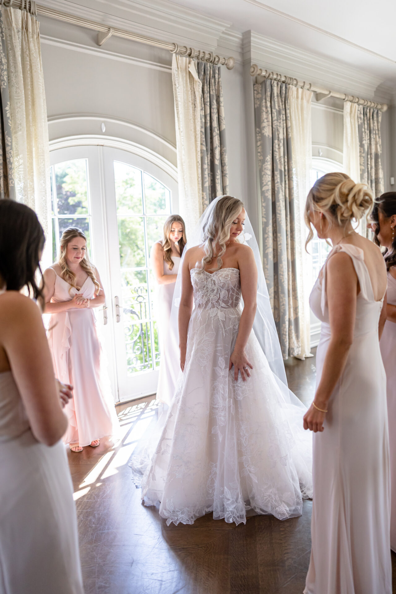 Bride with bridesmaids in sunlit room, preparing.