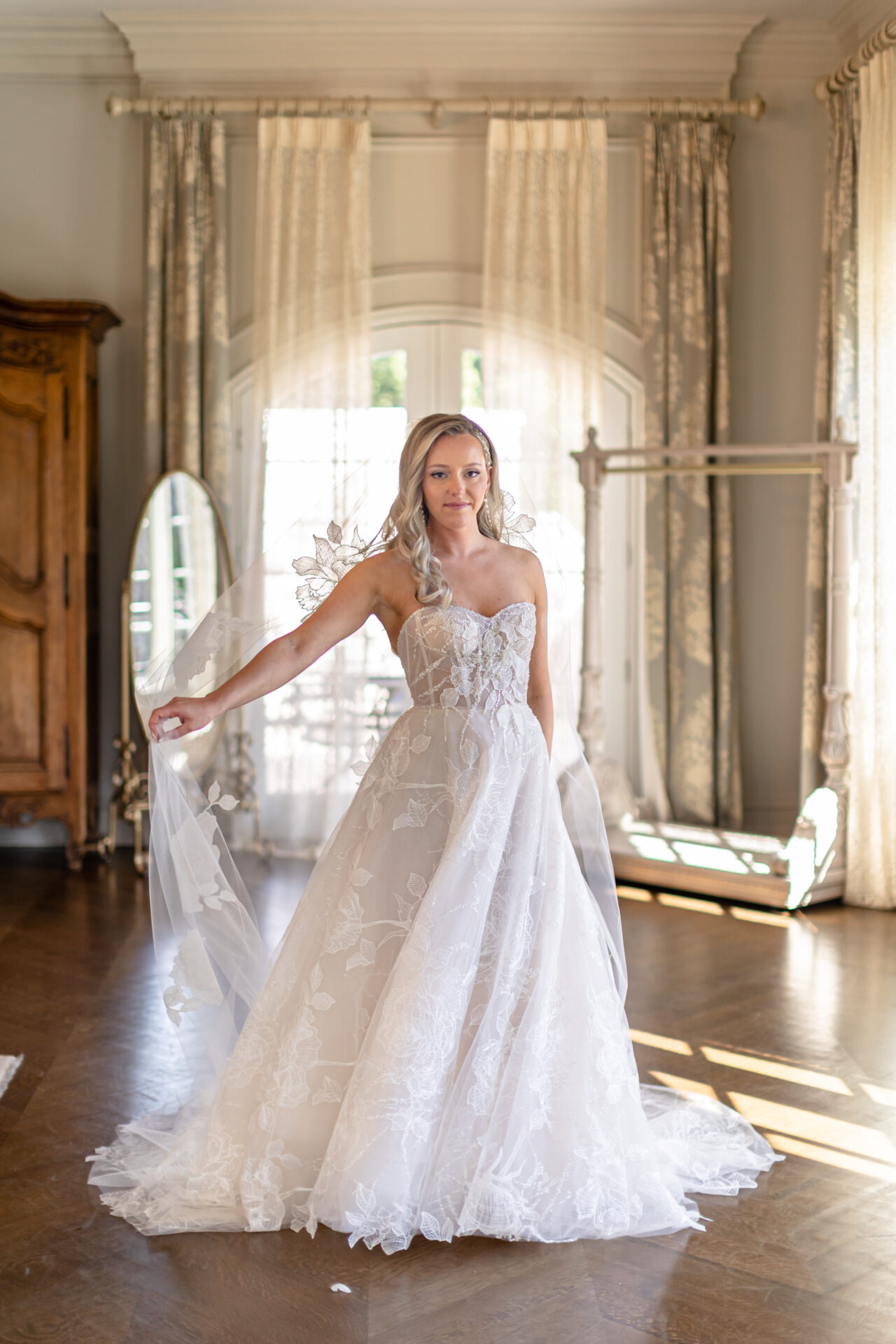 Bride in lace gown holds veil by sunlit windows.
